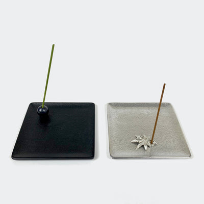 A black square incense holder tray on the left has a black ball incense holder with a green incense stick. A silver square incense holder tray on the right has a Japanese maple incense holder with a brown incense stick.