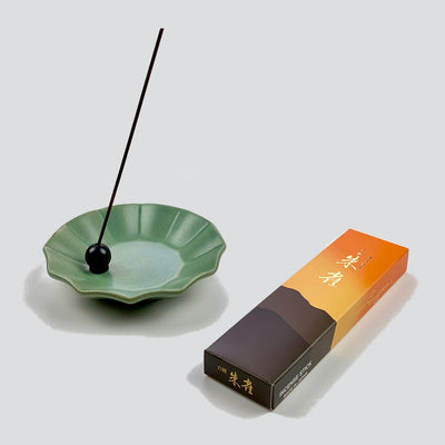 A stick of Byakudan Suzaku incense held with a black brass ball incense holder resting on a moss pleated dish with a box of Byakudan Suzaku lying in the front.