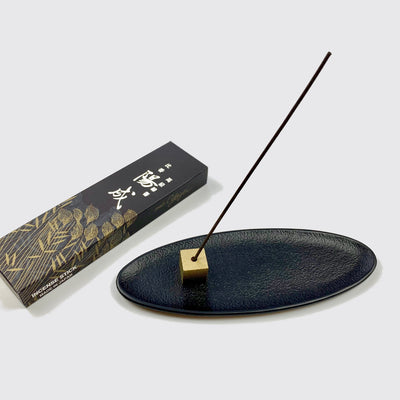 A stick of Jinko Yozei incense held with a gold brass cube incense holder resting on a black oval incense holder tray with a box of Jinko Yozei lying in the back.