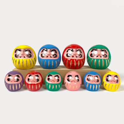 A full lineup of daruma dolls: medium daruma on top row (from left) yellow, blue, red, green; small daruma on bottom row (from left) purple, red, green, pink, blue, yellow.