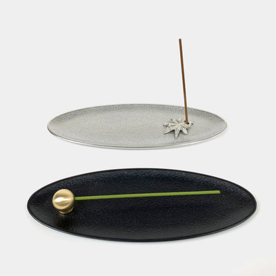 Silver oval incense holder tray in the back with a Japanese maple incense holder and a brown incense stick. Black oval incense holder tray in the front with a gold brass ball incense holder and a green incense stick.