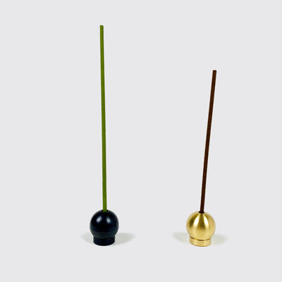 A black brass ball incense holder with a green incense stick on the left and a gold brass ball incense holder with a brown incense stick on the right.